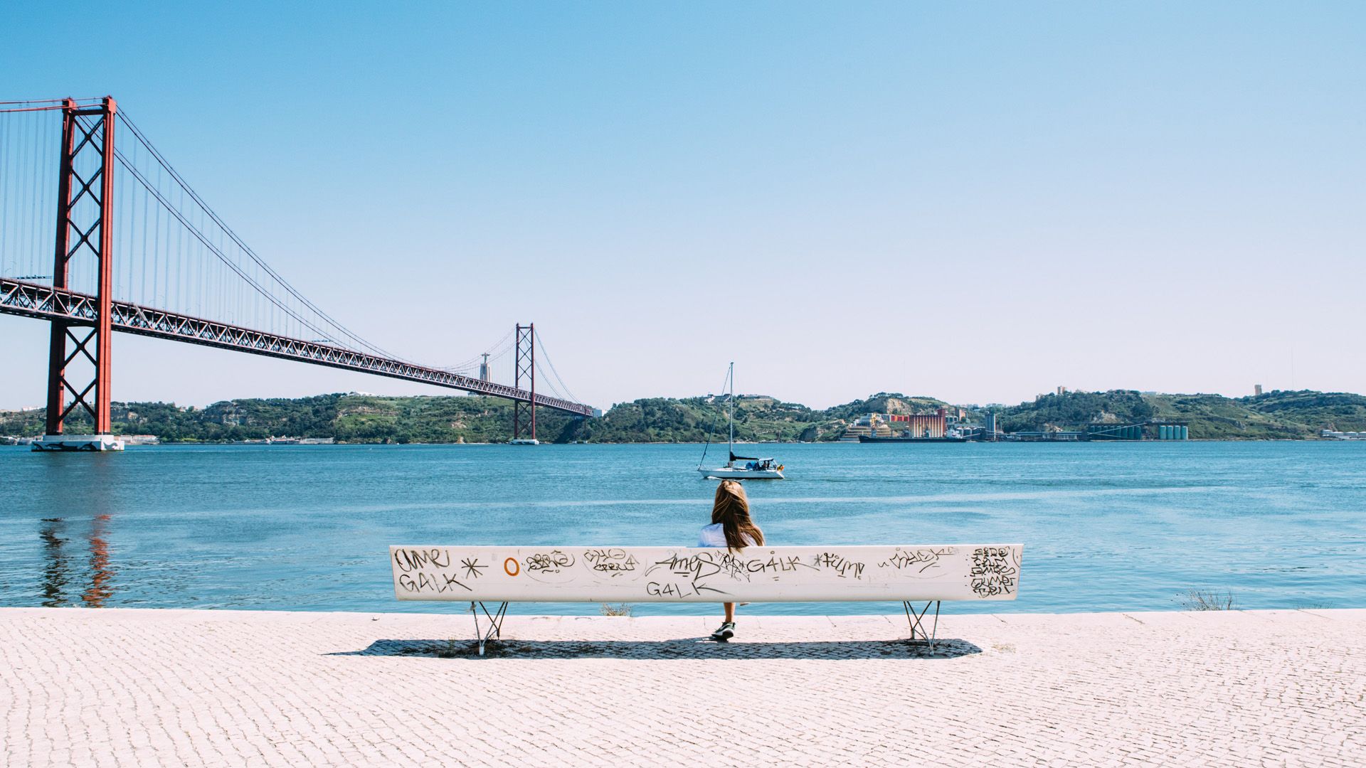 A woman is sitting on a bench overlooking Tagus River and Bridge 25th in the background.