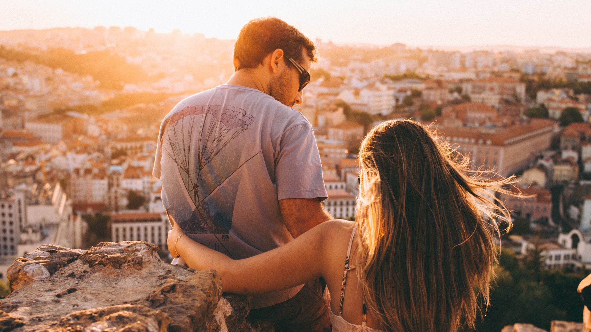 A man and a woman are sitting on top of a hill overlooking Lisbon.