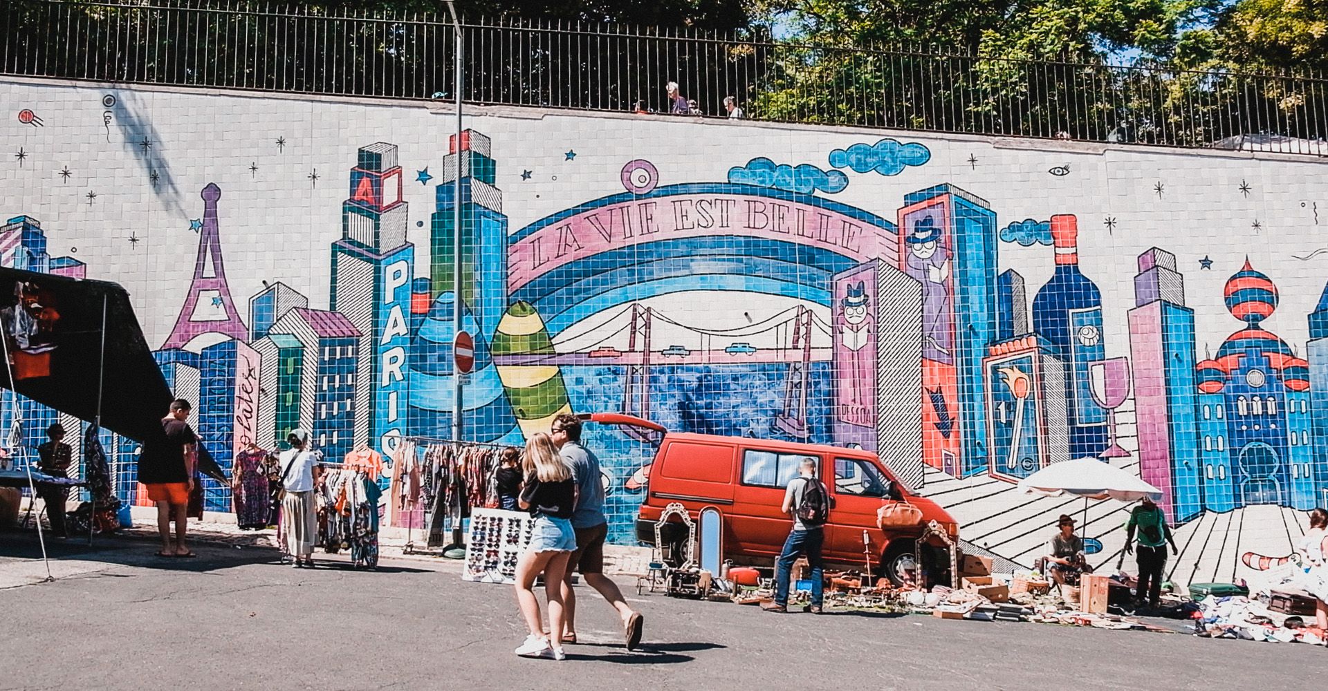 A group of people are walking in front of a mural on a wall near Flea Market in Lisbon.
