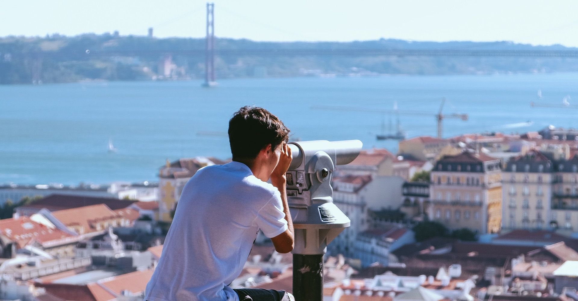 A boy is looking through binoculars (in São Jorge Castle) at a city overlooking Tejo River.