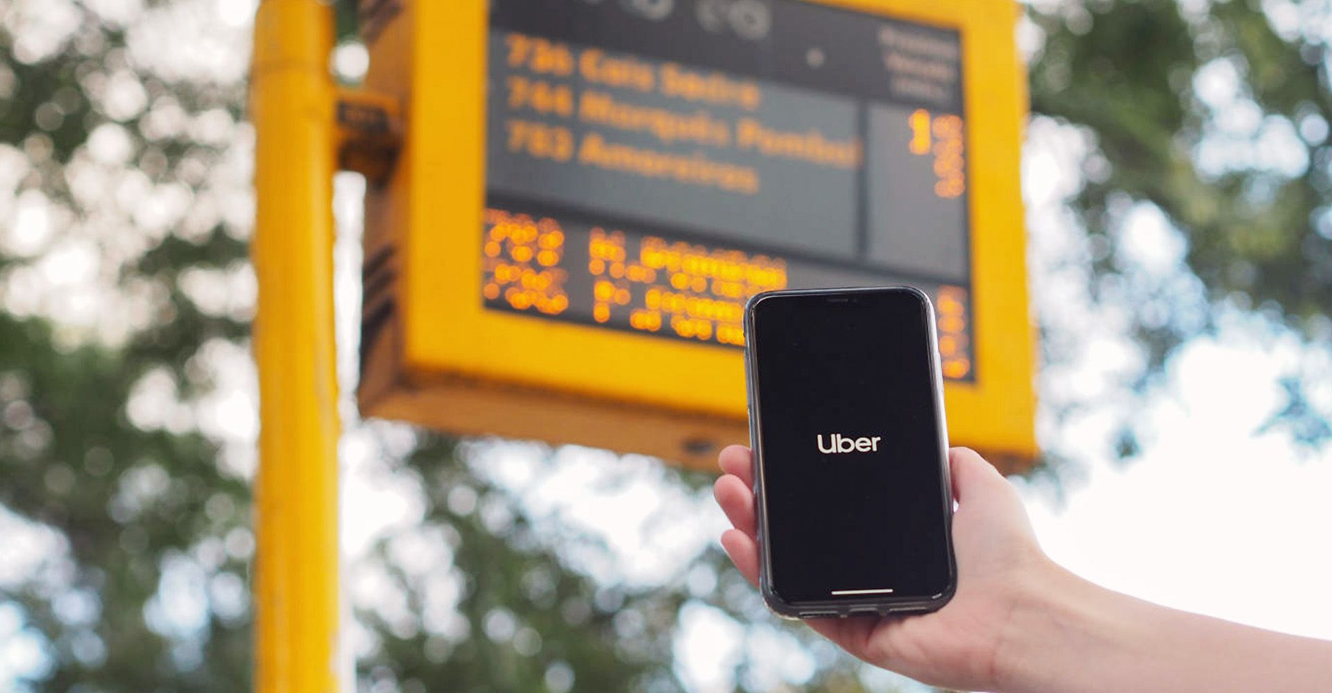 A person is holding a cell phone in front of a bus stop sign, with Uber app opening.